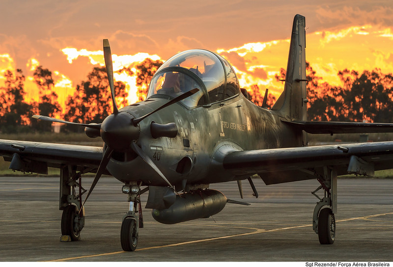 Aeronave A-29 Super Tucano em solo, com pôr do sol ao fundo destacando cores vibrantes.