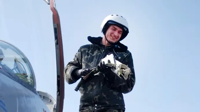 Piloto sorridente em um caça, segurando um item, com uma aeronave na parte de trás e um céu claro ao fundo.