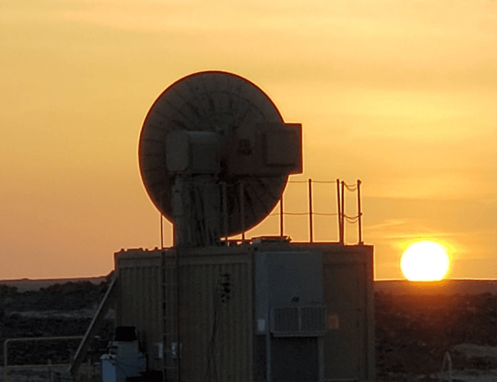 Sistema de armas de energia dirigida ao fundo, com o sol se pondo no horizonte, destacando a estrutura e os componentes técnicos.